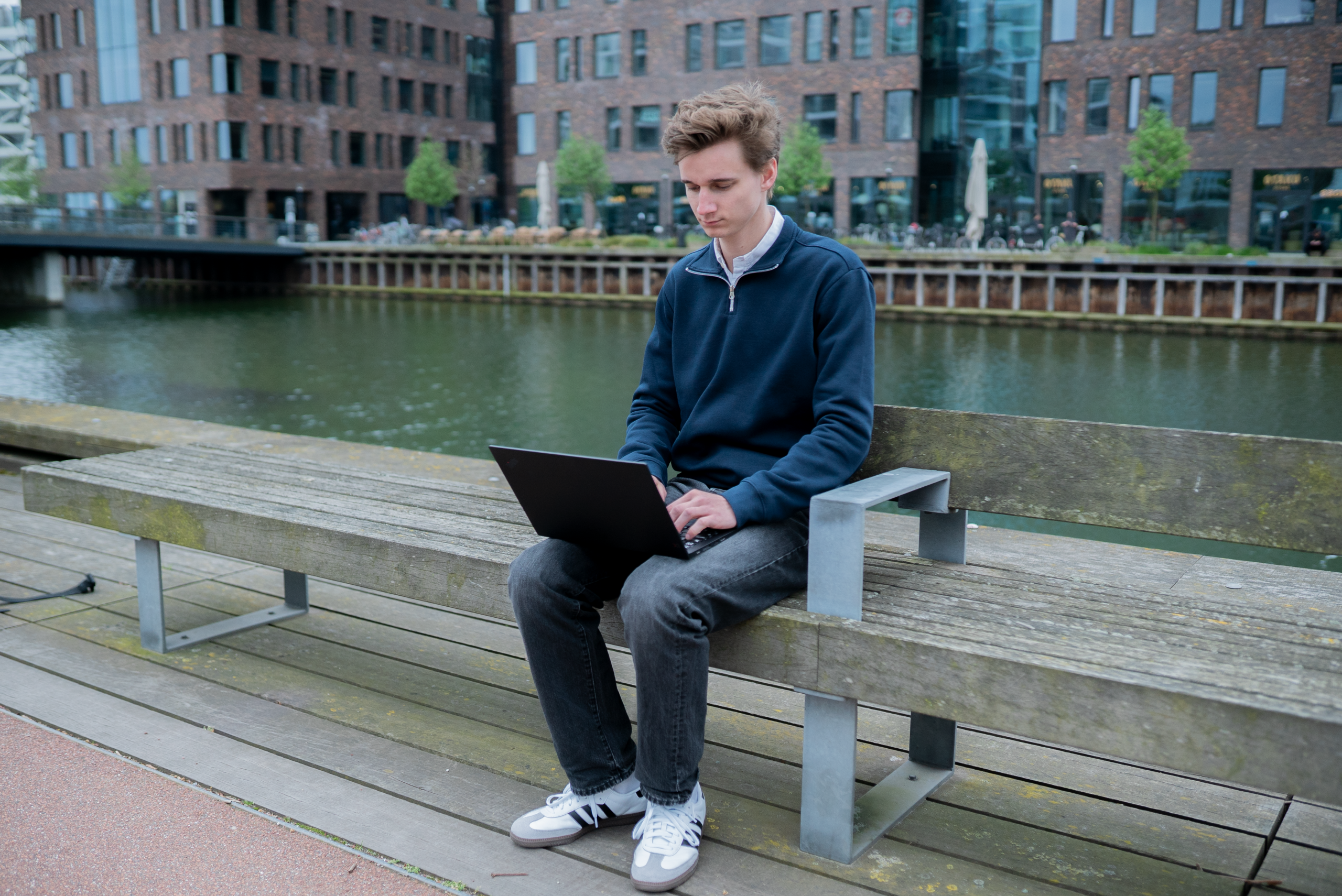 Ung mand arbejder på en Lenovo X1 Carbon-laptop, mens han sidder på en bænk ved en kanal i et moderne bymiljø