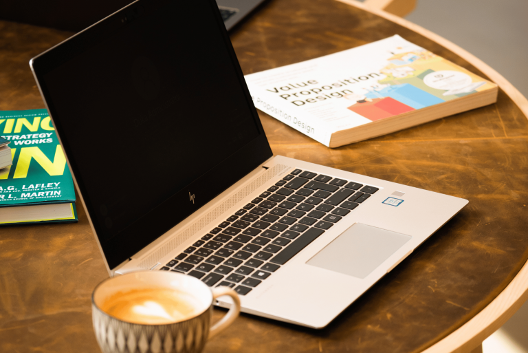 Sølvfarvet HP bærbar computer står åben på et bord ved siden af en kaffekop og bøger.