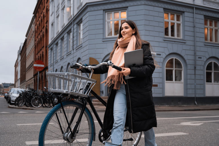 Kvinde går med sin cykel og en MacBook under armen i byen.
