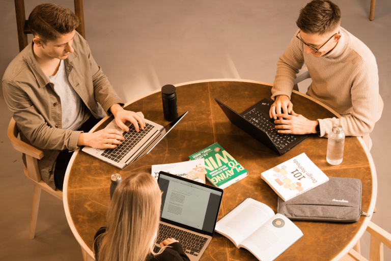Tre studerende arbejder på MacBook og PC omkring et bord med bøger og studiematerialer.